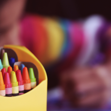 Värikkäässä kuvassa värikynälaatikko, taustalla lapsi piirtää.